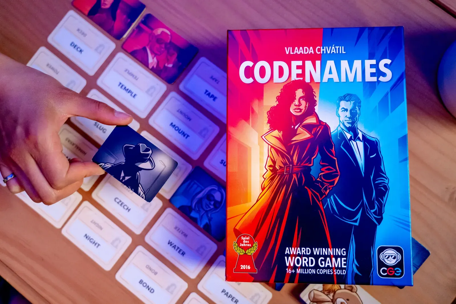 The Codenames box on a table, with a hand placing an agent card on the grid of word cards